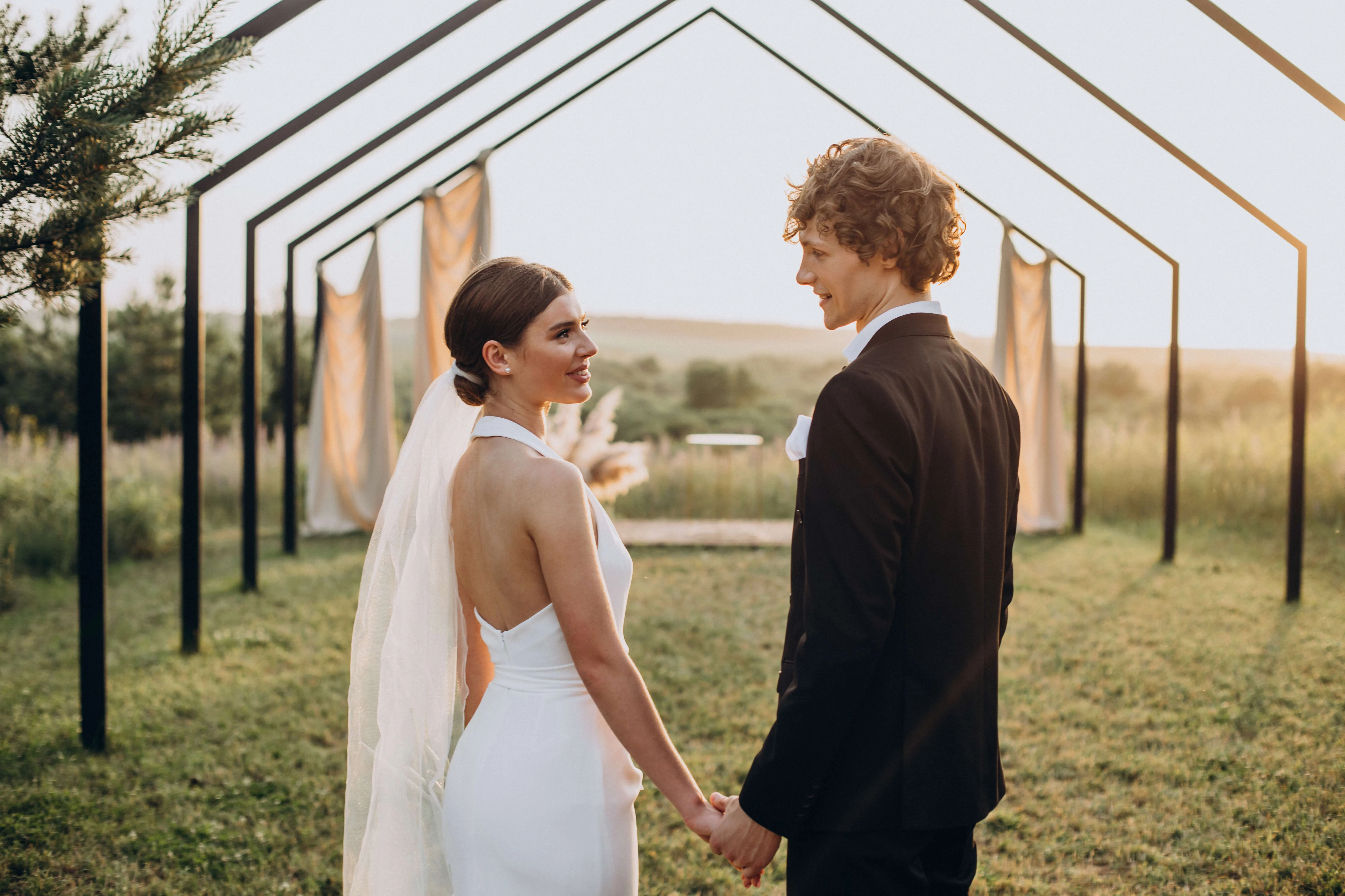 Bride,And,Groom,On,Their,Wedding,Ceremony Bride and groom holding hands and smiling at each other under a wooden arch structure at golden hour, captured by DTD Videography