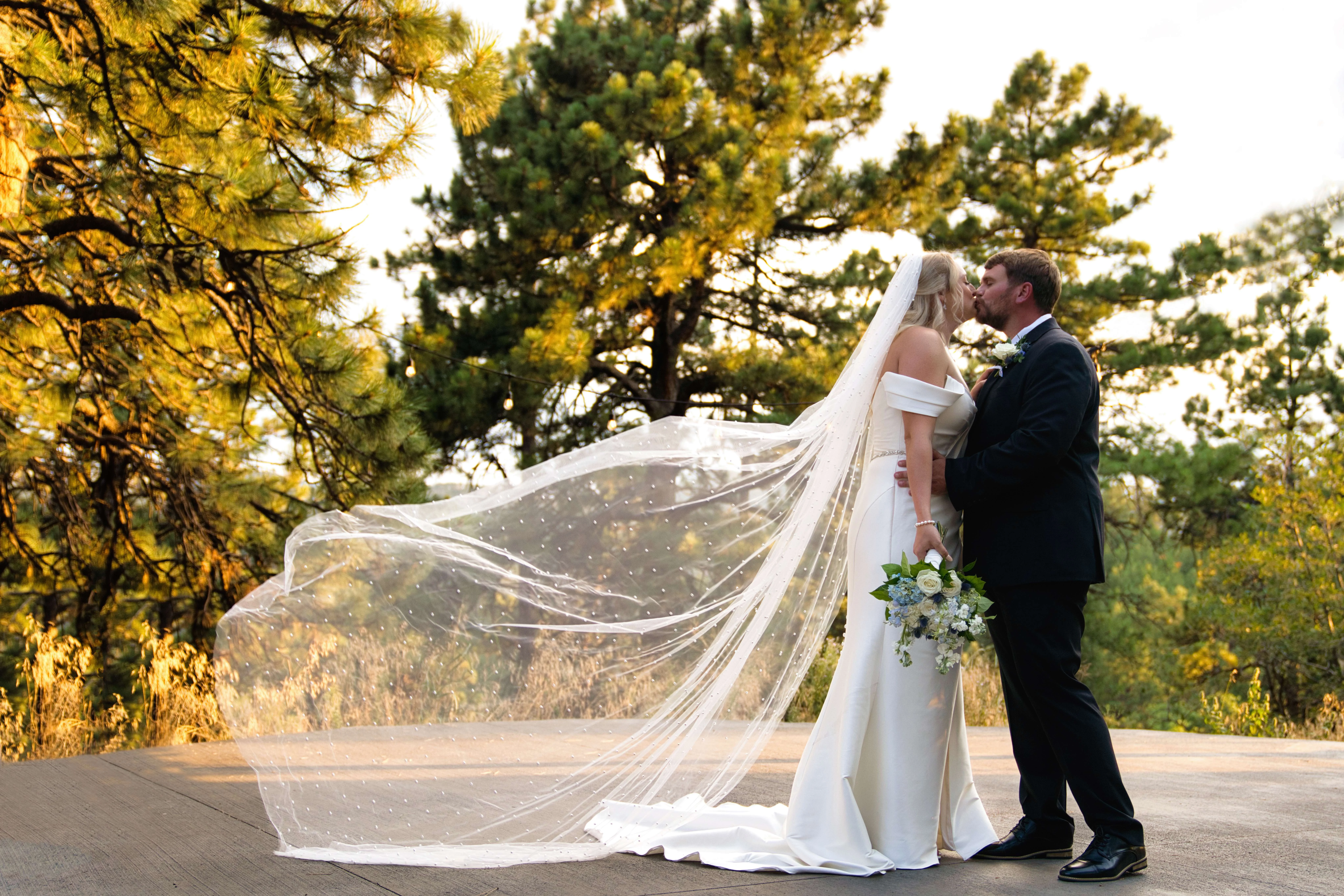 bride-groom-kiss-flowing-veil-wind-dtd-videography Bride and groom kissing outdoors with the bride's long veil dramatically flowing in the wind, surrounded by pine trees, captured by DTD Videography