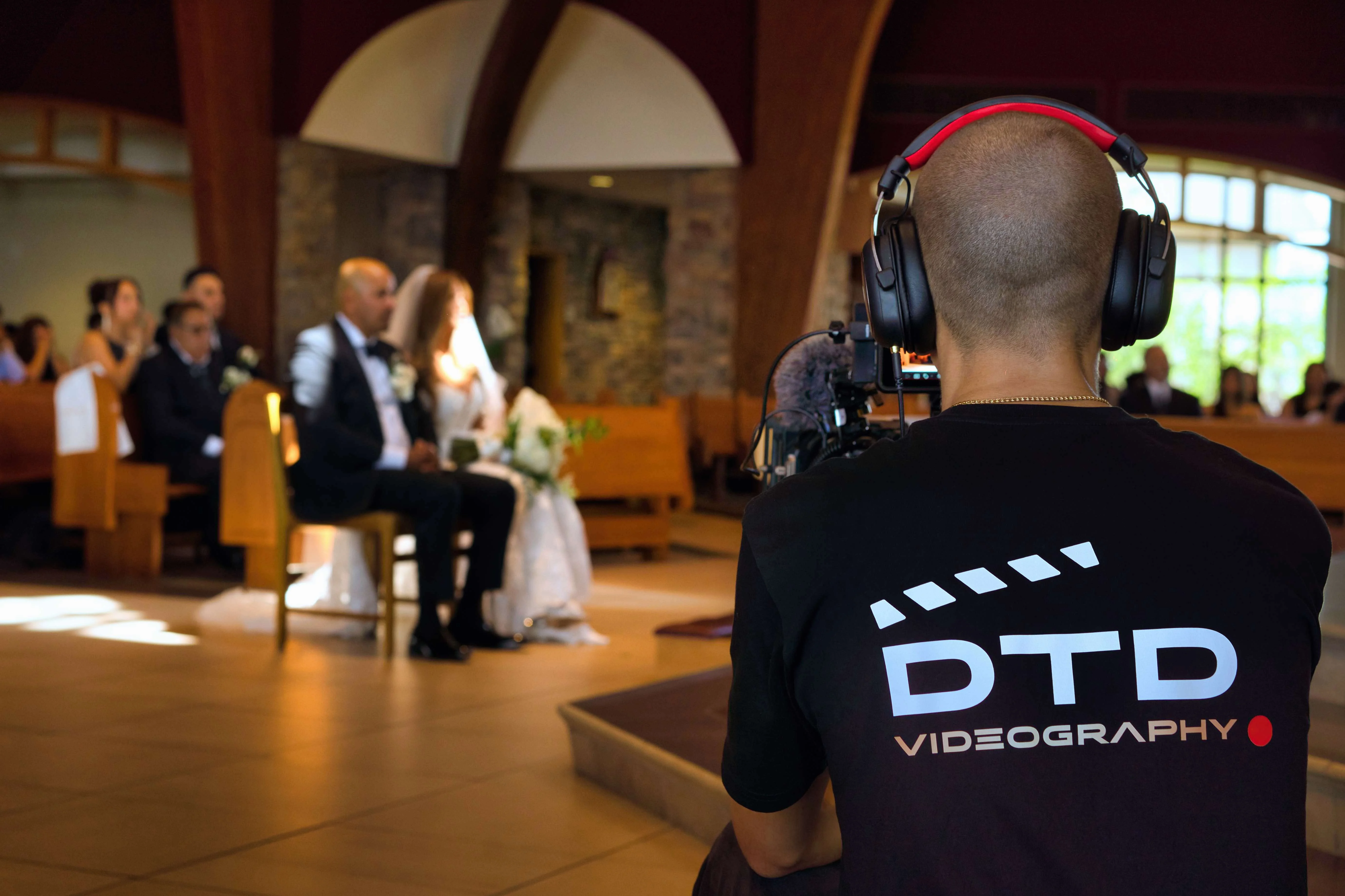 dtd-videography-filming-wedding-ceremony DTD Videography videographer filming a bride and groom seated during their wedding ceremony inside the church